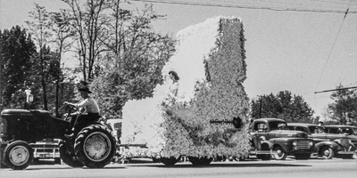 a tractor pulls a float in the shape of Idaho with a young woman seated on it