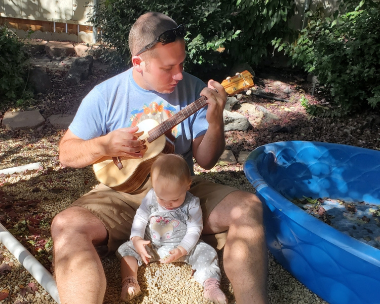 Image shows a man playing the ukulele to a toddler playing in the sand. 