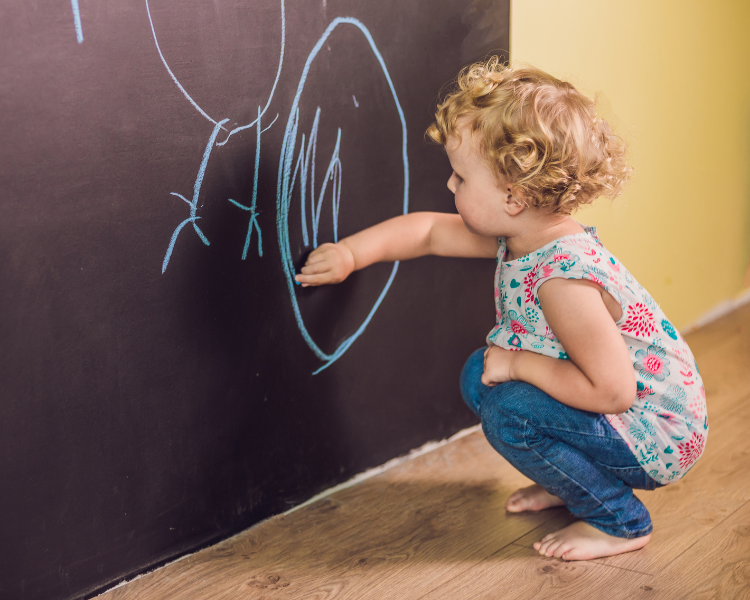 Image shows a toddler writing with chalk on a chalkboard wall.