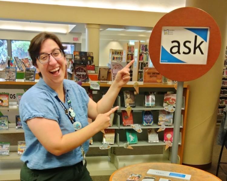 Librarian Irene smiles and points at a sign that says "ask"