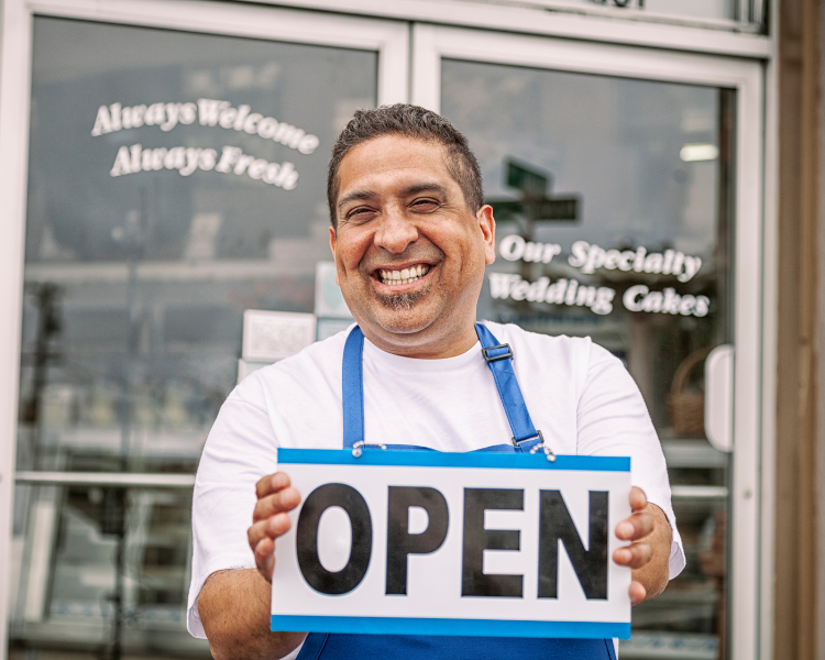 A smiling man holding an open sign in front of a business