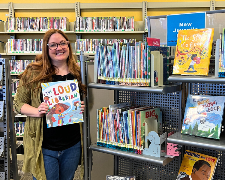Librarian holding a picture book in front of a shelf of new juvenile books.